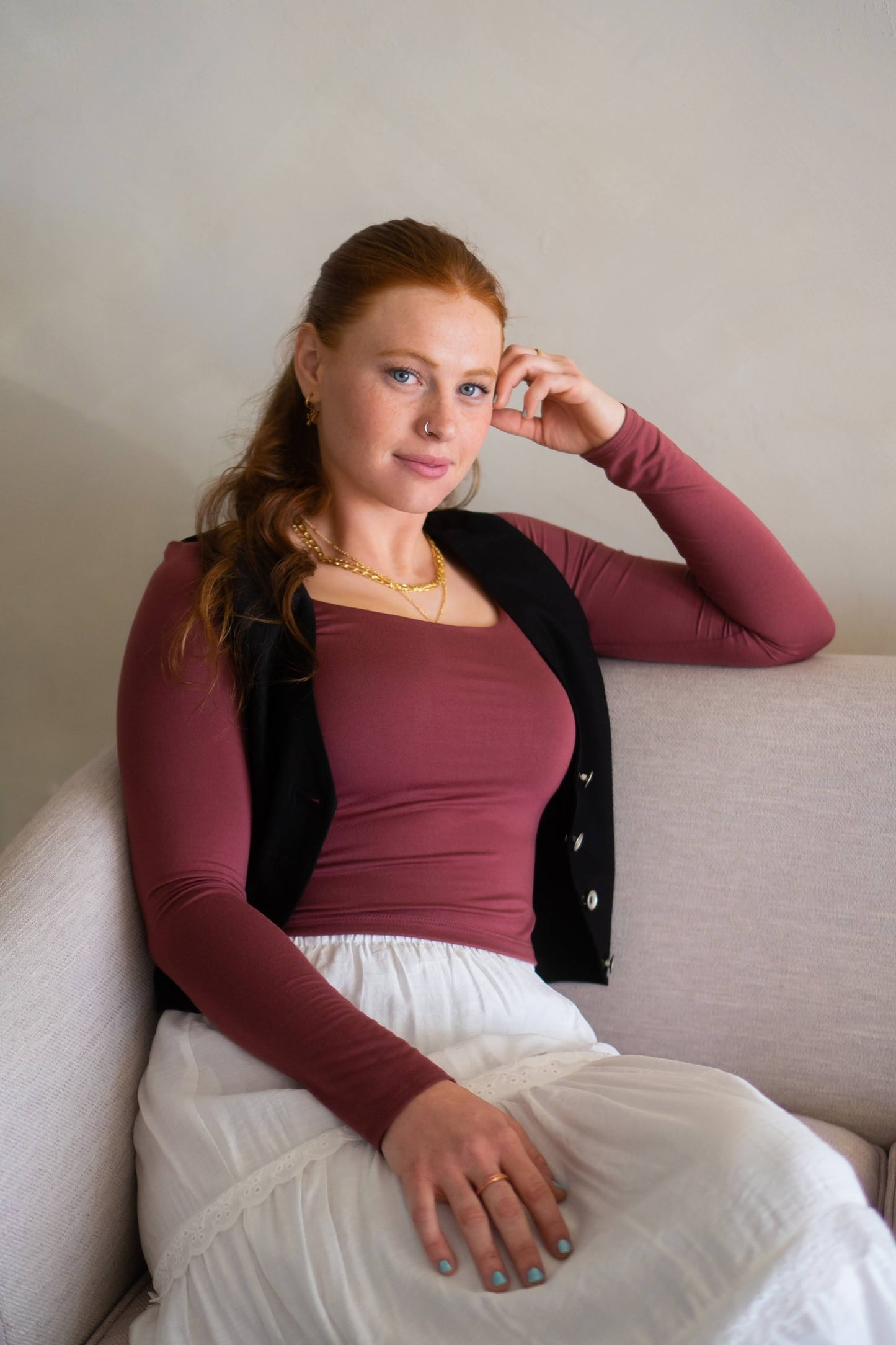 Woman sitting on a couch wearing a maroon top, with a black vest, and white skirt, with a neutral background.