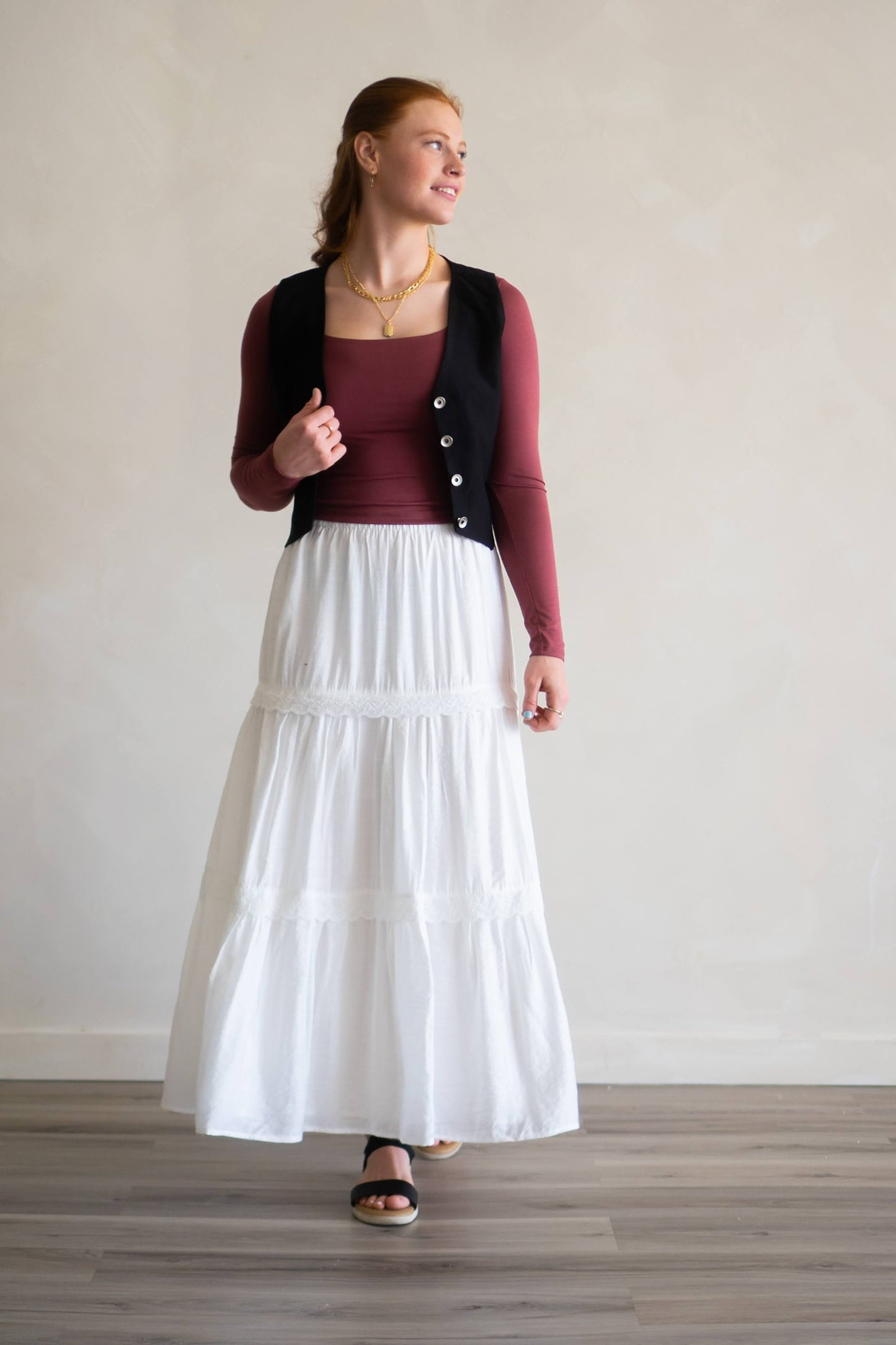 Woman wearing a white skirt, maroon top, and black vest against a plain background