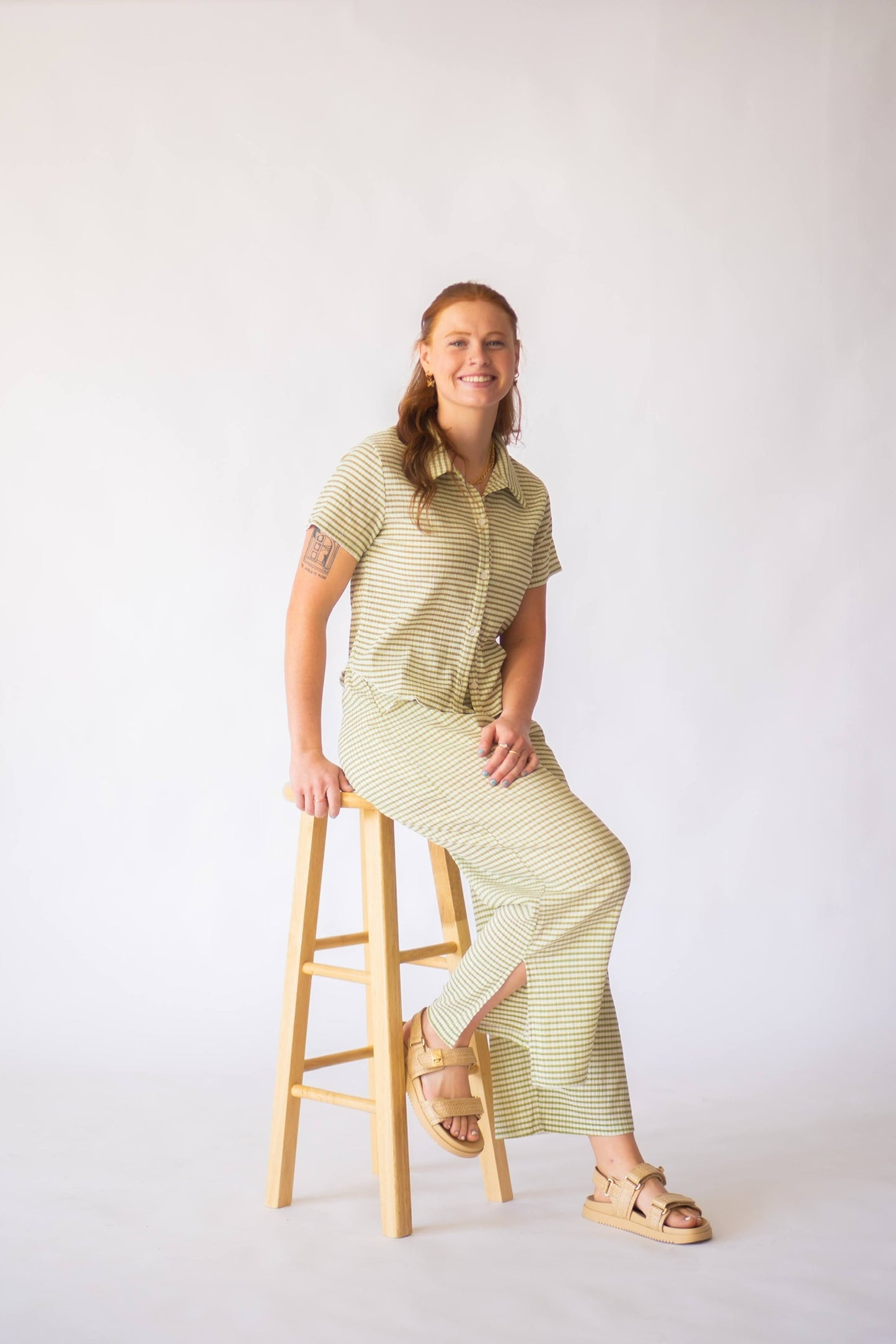 Woman in a green striped outfit sitting on a wooden stool against a white background