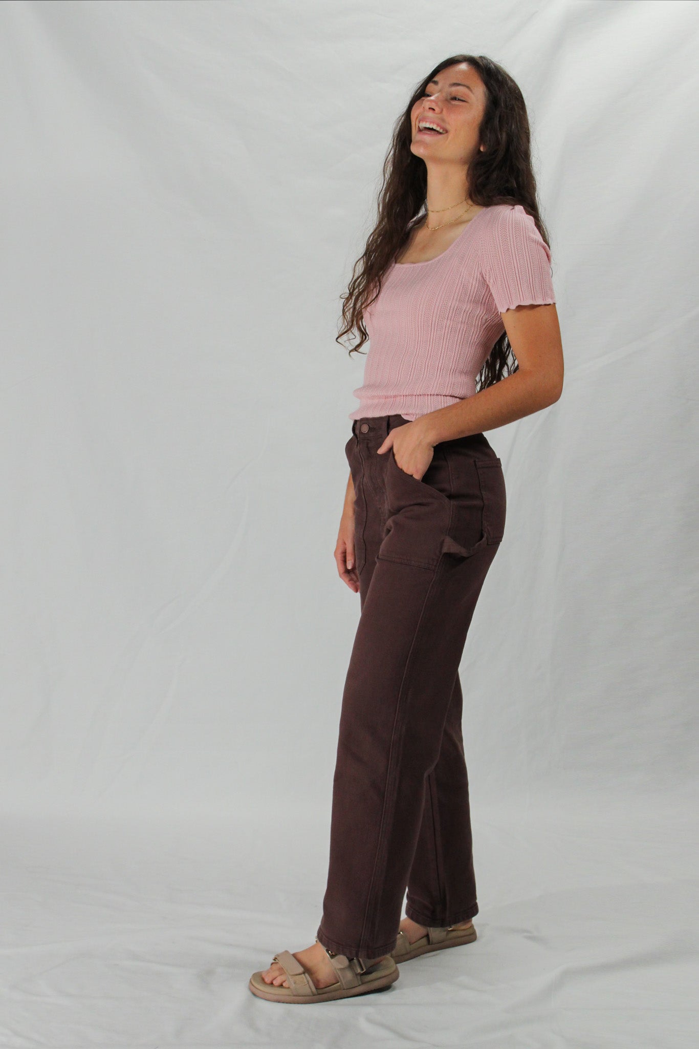 Woman wearing a pink top and brown carpenter pants against a white background