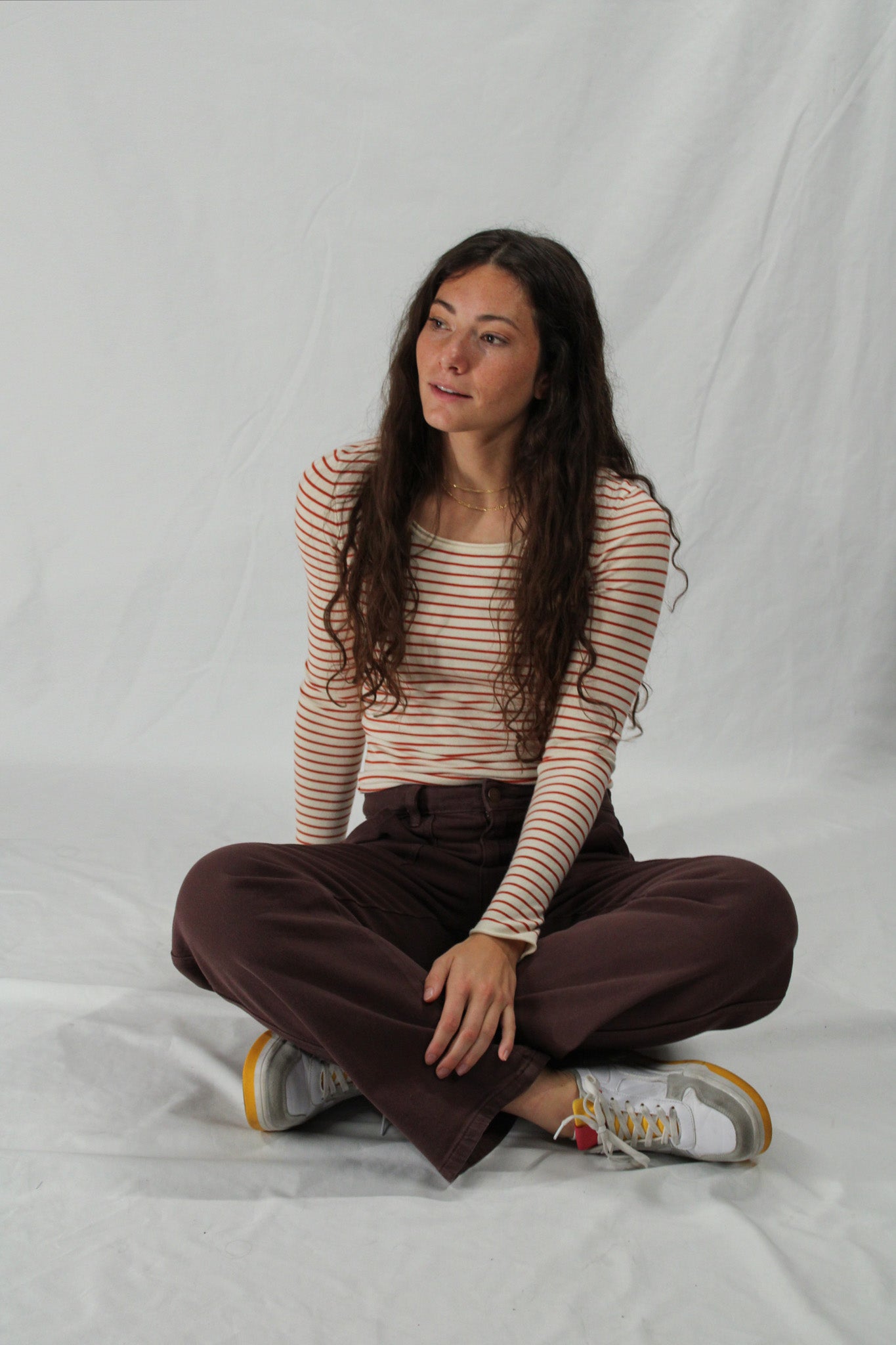 Woman sitting cross-legged on a white background wearing a striped shirt and brown pants.