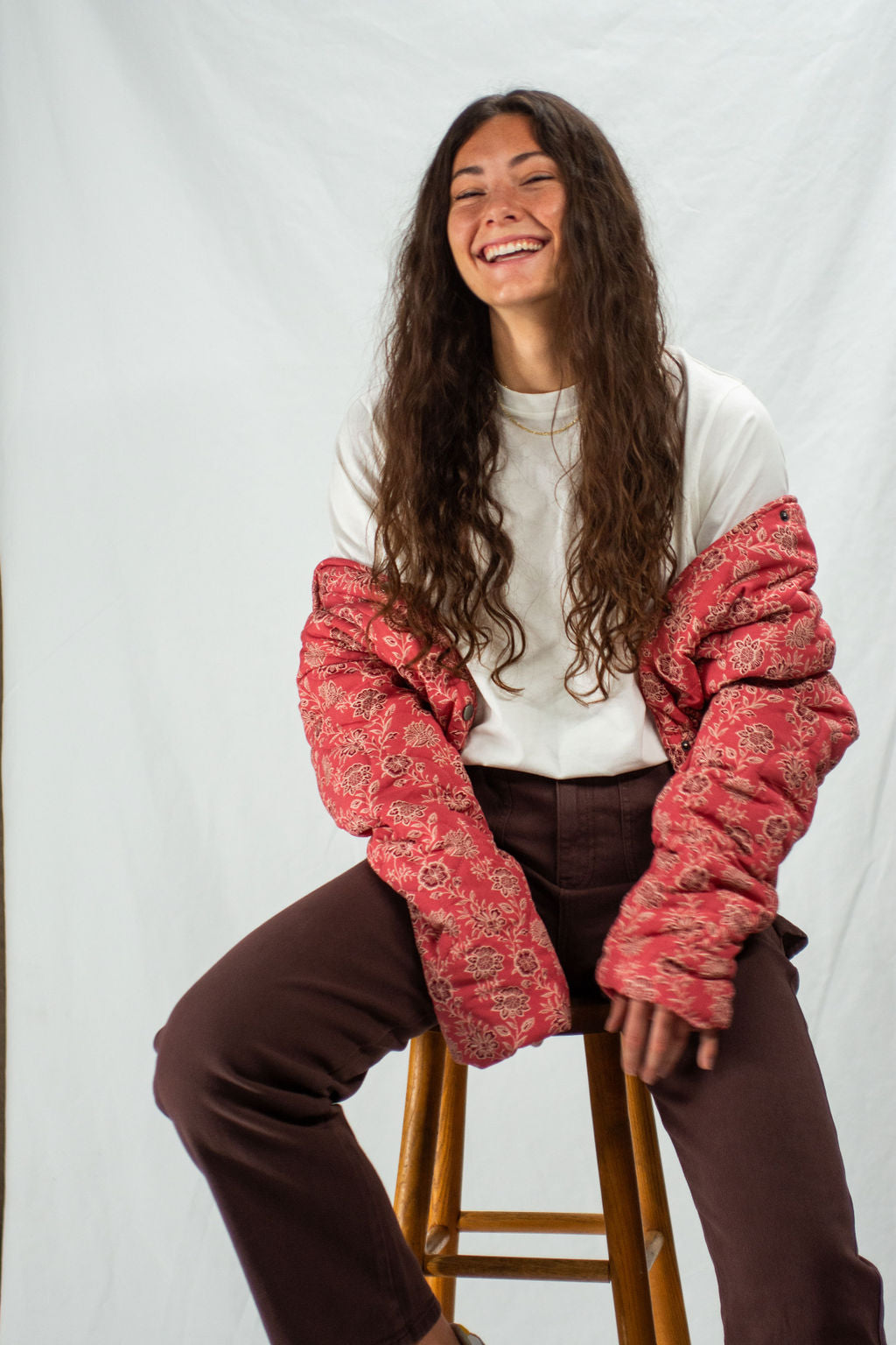Person wearing a red patterned quilted jacket over a white shirt and brown pants, sitting on a stool against a plain background.