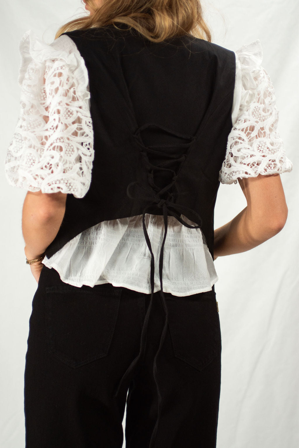 Person wearing a black vest over a white lace top with a plain background