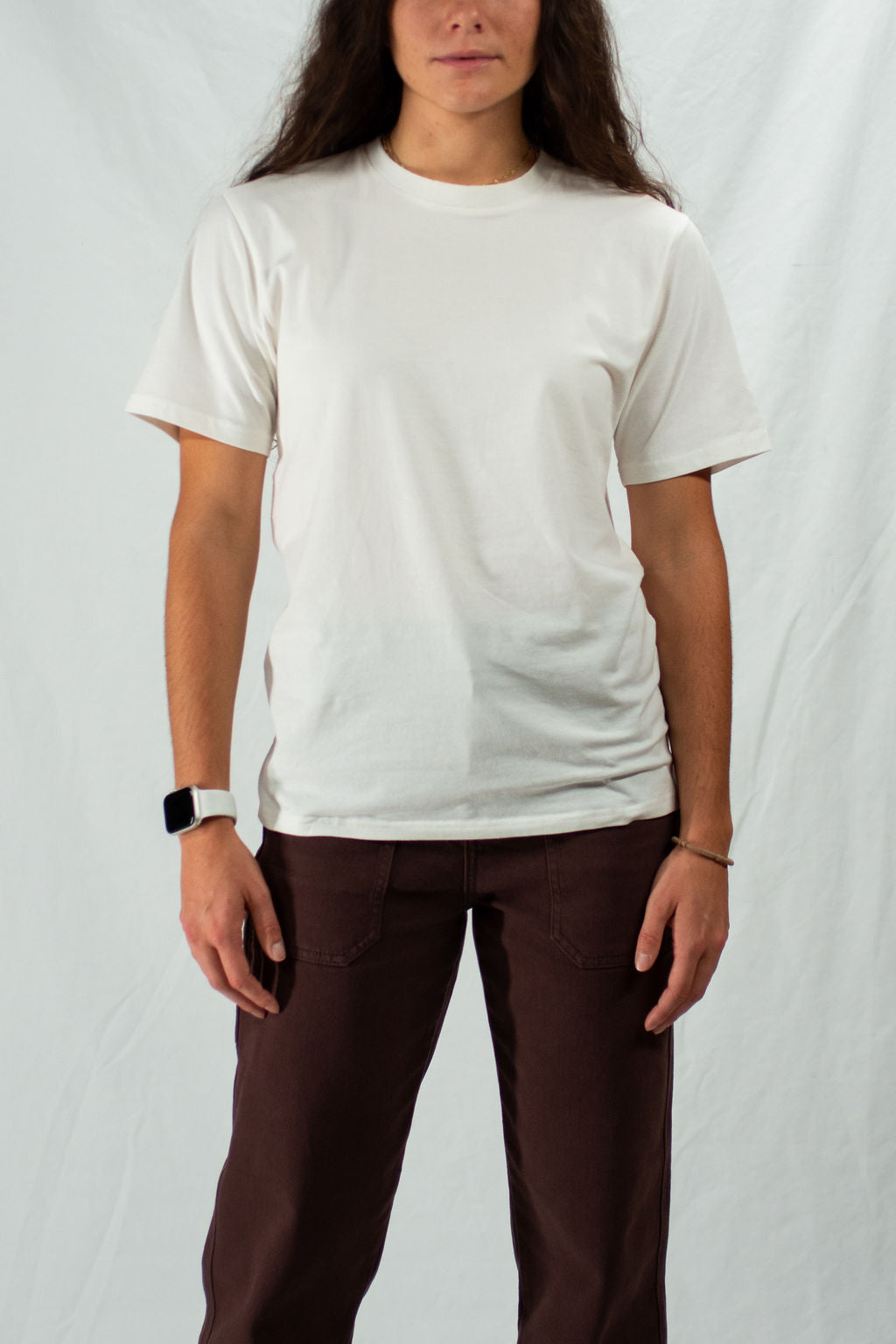 Woman wearing a plain white t-shirt and brown pants against a plain background.