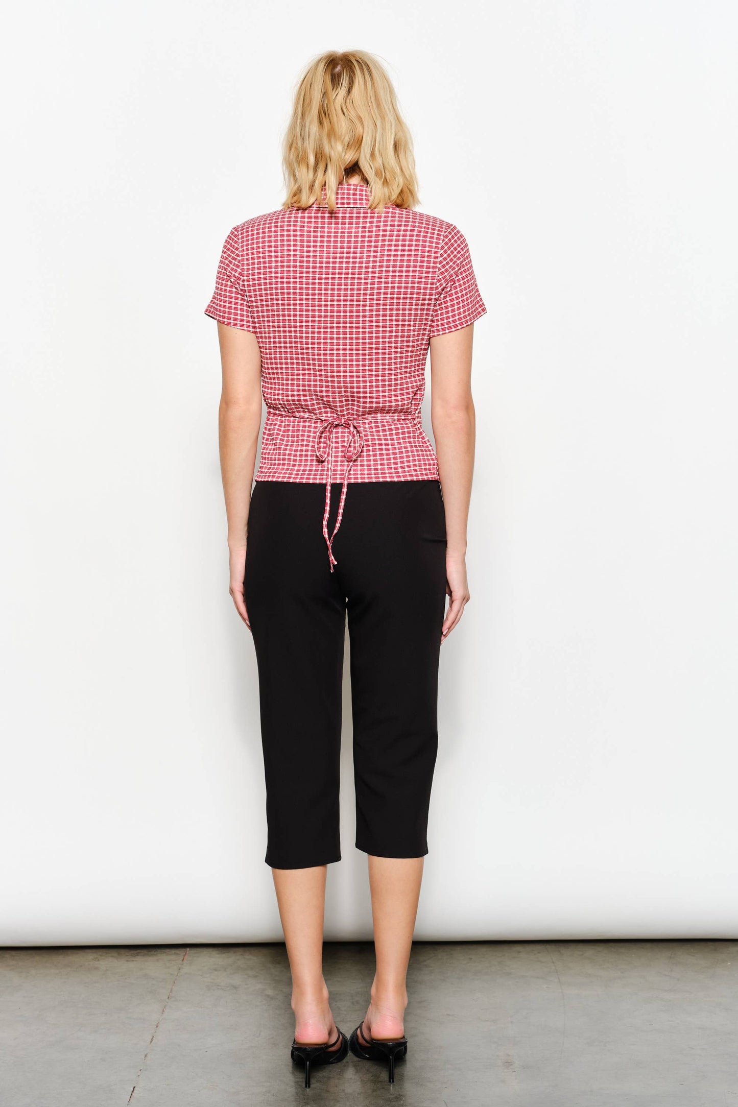 Person wearing a red checkered shirt and black pants against a white background, view from behind, the top features a tie back