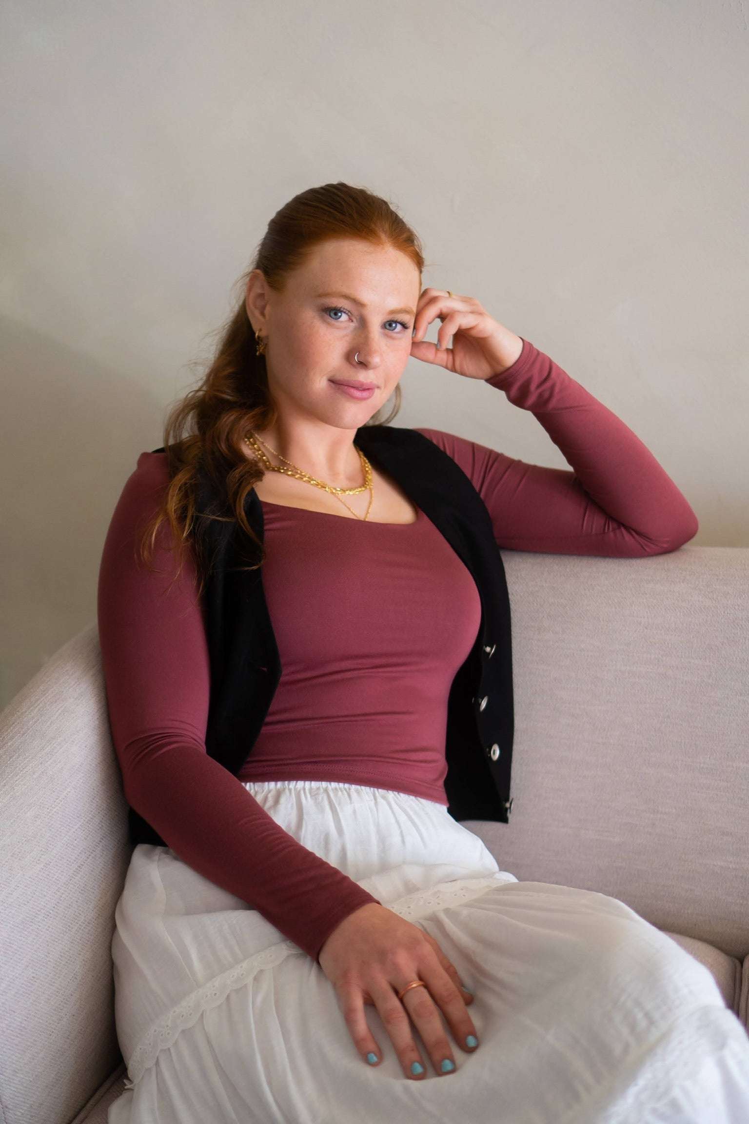Woman sitting on a couch wearing a maroon top, with a black vest, and white skirt, with a neutral background.