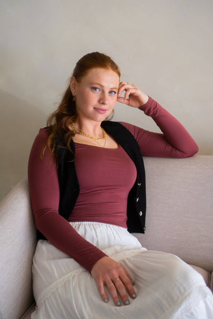 Woman sitting on a couch wearing a maroon top, with a black vest, and white skirt, with a neutral background.