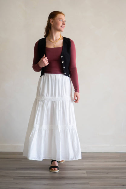Woman wearing a white skirt, maroon top, and black vest against a plain background
