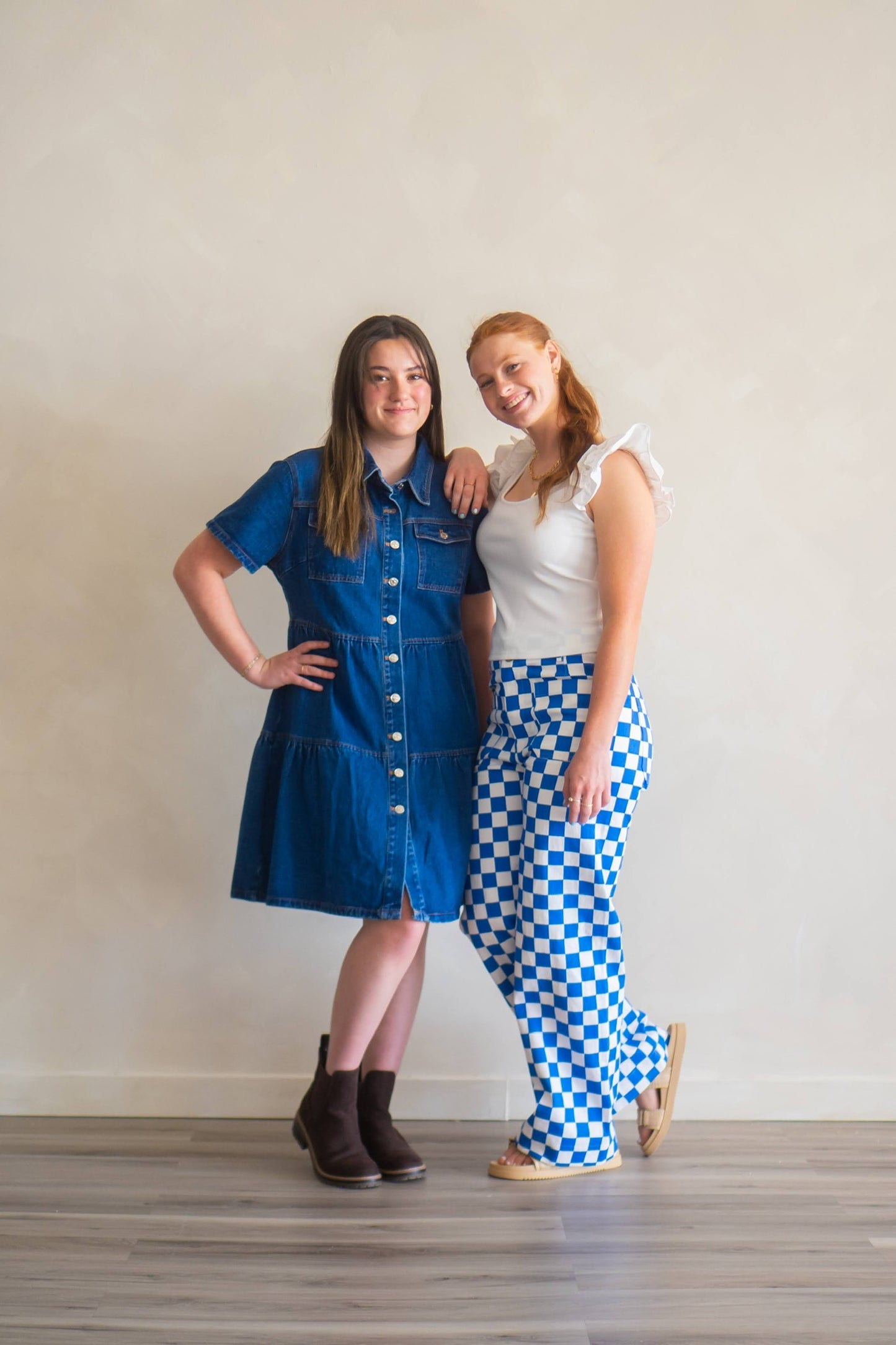 Two woman smiling at camera, left woman wears a denim dress that's knee length, other is wearing blue and white checkered pants, and white blouse with flutter sleeves.