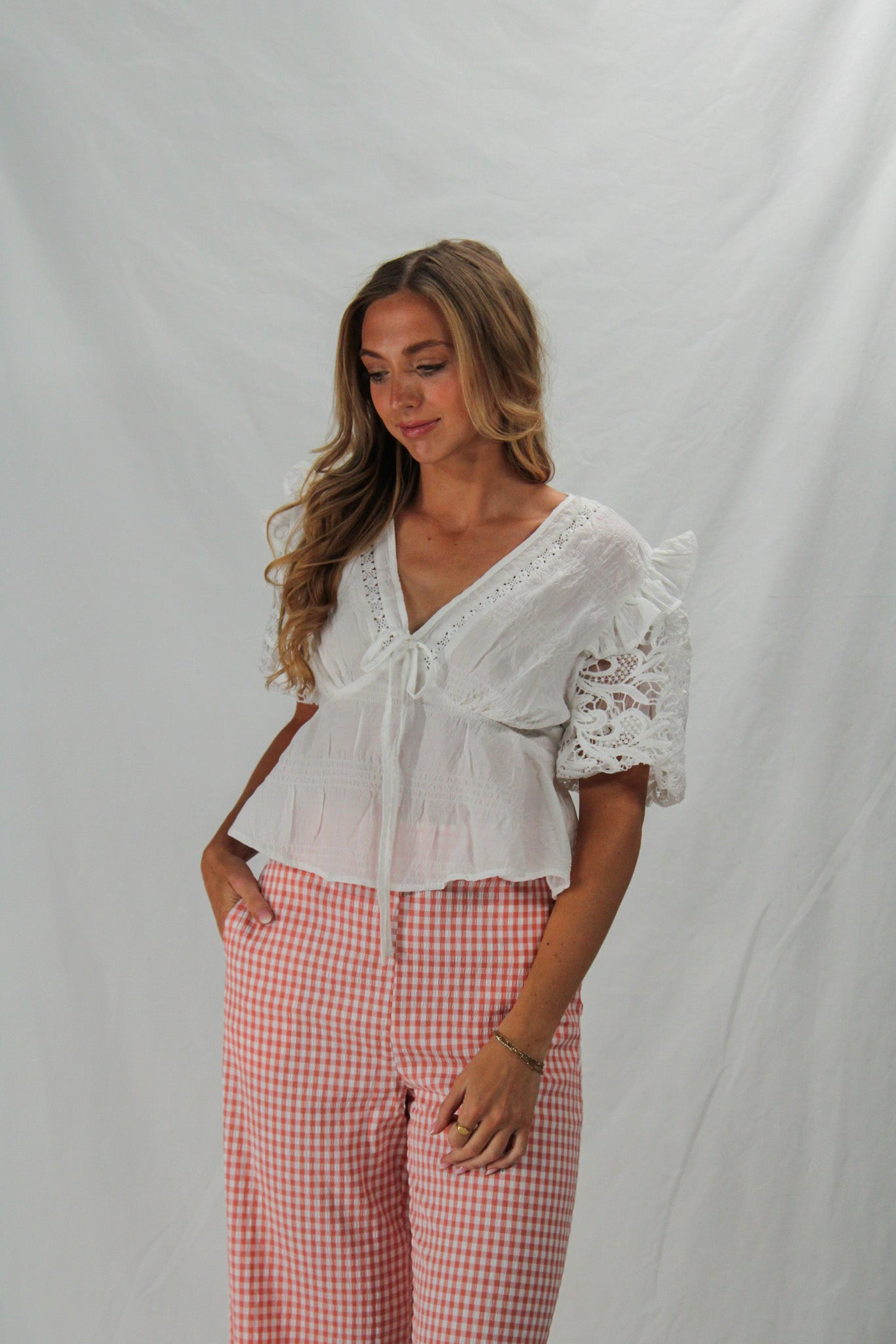 Woman wearing a white lace top and pink checkered pants against a plain background