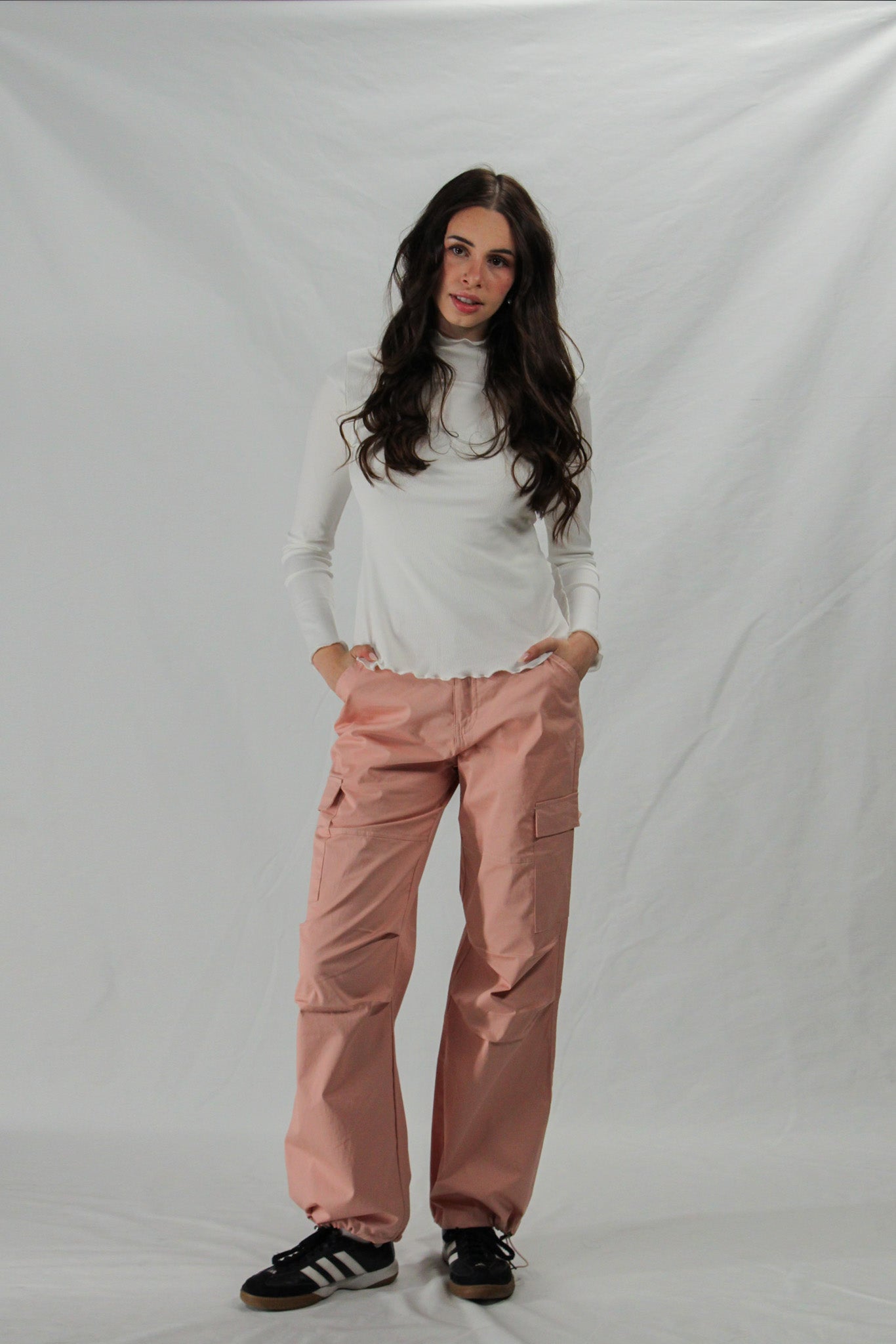 Woman wearing a white lettuce hem turtleneck and pink cargo pants on a plain background