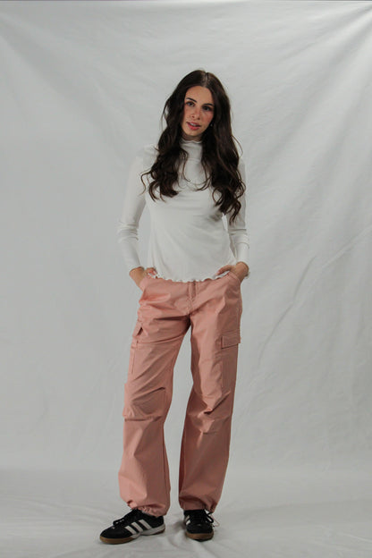 Woman wearing a white lettuce hem turtleneck and pink cargo pants on a plain background