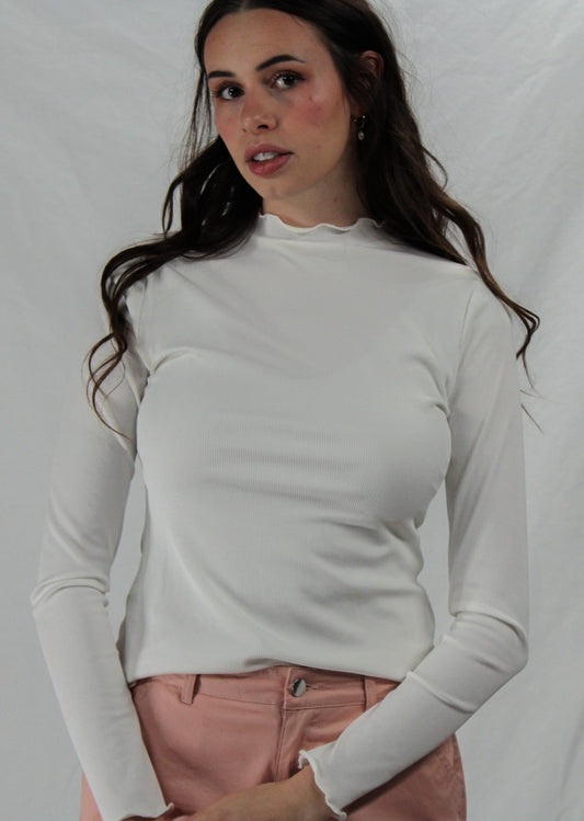 Woman wearing a white long-sleeve lettuce hem top and pink pants against a plain background