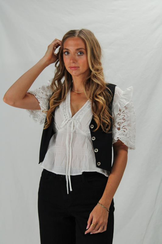 Woman wearing a white blouse with lace sleeves and a black vest against a plain background