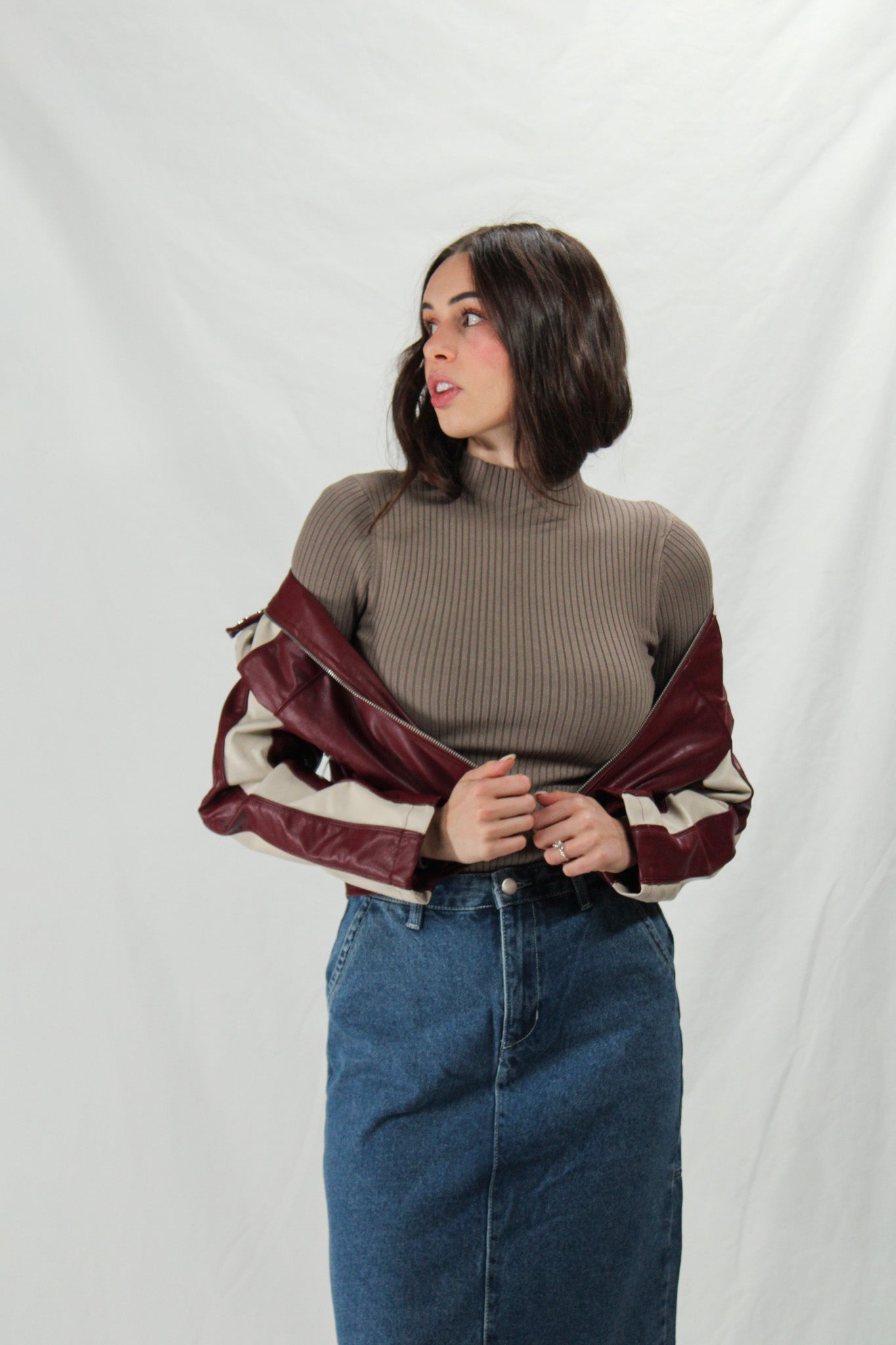 Woman wearing a brown ribbed sweater and blue jeans, with a maroon jacket with white lining draped over her arms.