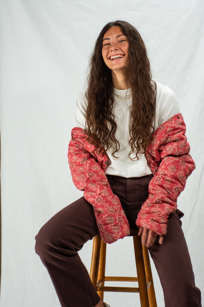 Person wearing a red patterned quilted jacket over a white shirt and brown pants, sitting on a stool against a plain background.