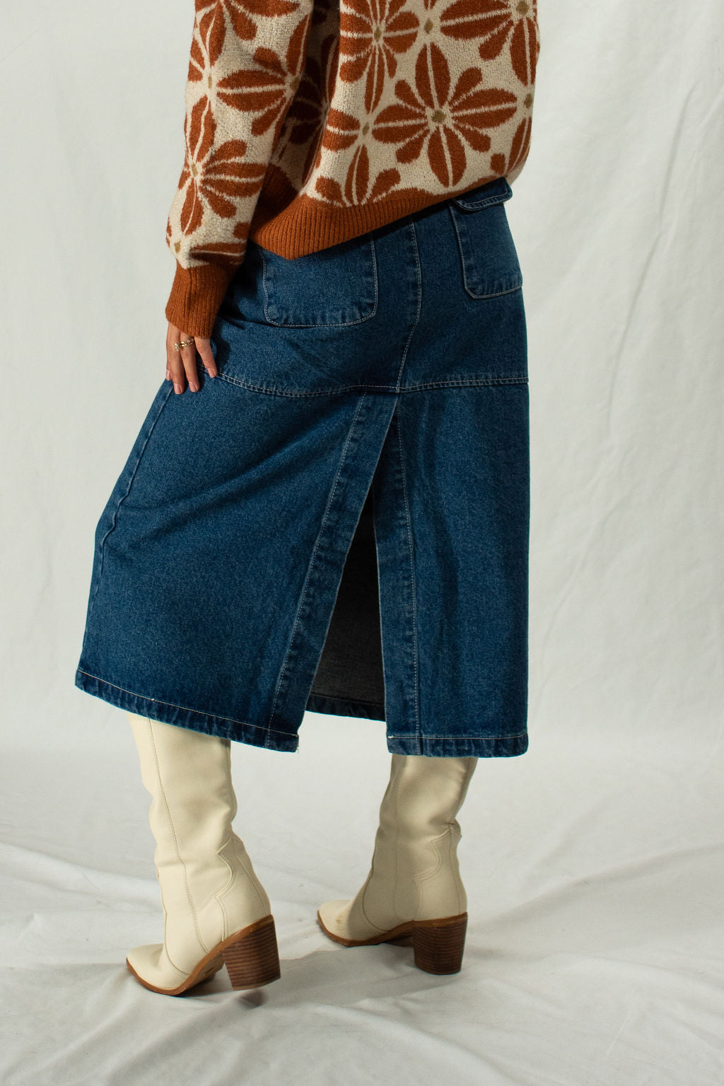 Person wearing a floral sweater, blue denim skirt with a slit in the back, and white boots on a plain background