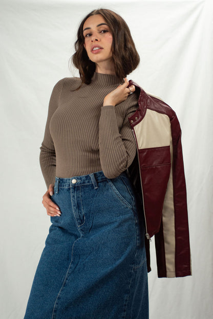 Woman wearing a brown ribbed sweater and blue jeans, holding a maroon and cream leather jacket.